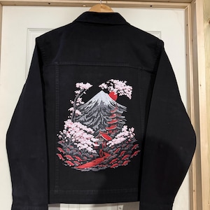 Può includere: Giacca di jeans nera con un design ricamato dettagliato sul retro. Il design presenta una montagna, una pagoda e fiori di ciliegio nei toni del grigio, rosso e rosa, su uno sfondo nero. La giacca ha un colletto classico e chiusura con bottoni.