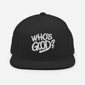 May include: A black baseball cap with a flat brim and a structured crown. The front of the cap features white embroidered text that reads "WHO'S GOOD?" in a bold, slightly slanted, and stylised font with a clean, modern appearance.
