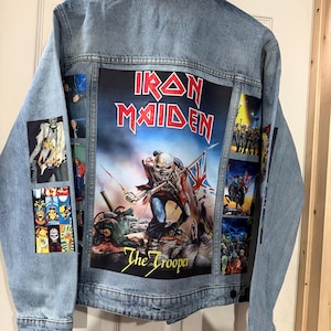 May include: A light blue denim jacket with a large Iron Maiden graphic on the back, featuring the text "The Trooper." The sleeves have additional graphics. The jacket is hanging on a wooden door.