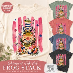 May include: A short-sleeved t-shirt featuring a graphic of three stacked frogs. The top frog wears a blue and white striped shirt, the middle frog has a floral pattern, and the bottom frog has colourful abstract shapes. Pink brushstrokes, flowers, and stars surround the frogs.