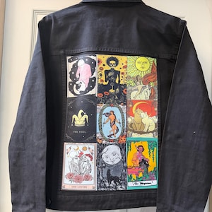 May include: A black denim jacket featuring a large rectangular back panel printed with a grid of nine colourful tarot card illustrations, including depictions of skeletons, celestial bodies, cats, and figures with the text The Fool, The Lovers, and The Magician.