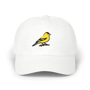 Puede incluir: Una gorra de béisbol blanca con un gráfico central de un jilguero amarillo con detalles de alas en negro y blanco posado sobre una pequeña rama marrón. La gorra tiene una corona estructurada, una visera curva y un botón en la parte superior.