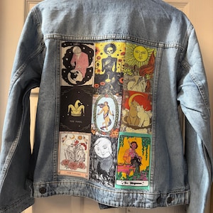 May include: Pale blue denim jacket adorned with tarot card patches on the back. The patches display colourful illustrations of tarot cards, including "The Lovers" and "The Magician". The jacket has a traditional button-up design.