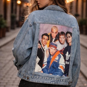 Peut inclure: Veste en jean bleu clair avec un graphisme coloré au dos représentant un groupe de personnes. La veste a un design classique avec des boutons et des poches. L'image est placée sur un arrière-plan flou.
