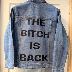 Könnte beinhalten: Hellblaue Jeansjacke mit dem Aufdruck "THE BITCH IS BACK" in großen schwarzen Buchstaben auf dem Rücken. Die Jacke hängt an einem schwarzen Kleiderbügel vor einer weißen Tür.