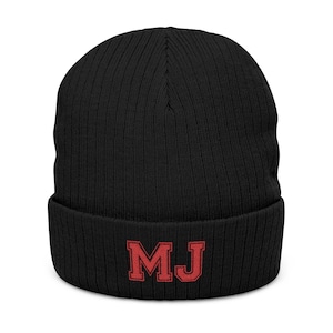 Op de afbeelding: Een zwarte, geribbelde gebreide muts met een omgeslagen boord. Op de voorzijde van de boord zijn de letters MJ geborduurd in een opvallend rood lettertype met een donkerrode omlijning, in een klassieke universitaire blokstijl.