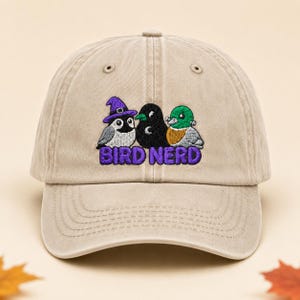 Könnte beinhalten: Eine beige Baseballkappe mit einer Stickerei von drei Vögeln: einer trägt einen violetten Hexenhut, einer ist schwarz und einer ist eine Stockente. Unter den Vögeln ist der Text BIRD NERD in fetten violetten Buchstaben auf die Vorderseite gestickt.
