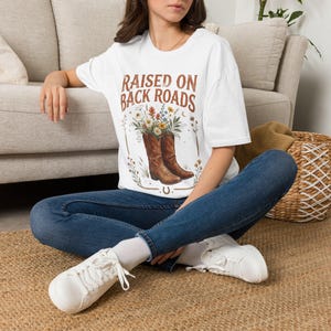 Op de afbeelding: Een wit T-shirt met korte mouwen en een afbeelding van bruine cowboylaarzen gevuld met kleurrijke wilde bloemen. De tekst "RAISED ON BACK ROADS" staat in bruin boven de laarzen, met kleine bloemaccenten en een hoefijzer eronder.