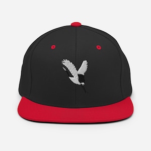 May include: A black baseball cap with a vibrant red flat brim and a red top button. The front panel features an embroidered white and black crane in flight. The cap includes red eyelets on the crown, providing a distinct contrast to the black fabric.