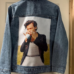 May include: A light blue denim jacket featuring a large printed image on the back. The image is a portrait of a person wearing a black suit and white dress. The jacket is suspended from a wooden hanger.