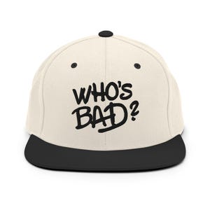 May include: A cream-coloured flat-brimmed baseball cap with a black brim and a black button on top. The phrase "WHO'S BAD?" is printed in bold, black letters across the front of the cap.