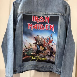 May include: Light blue denim jacket with a large graphic on the back featuring the band name "Iron Maiden" in red and the album art for "The Trooper". The artwork depicts a skeletal figure in battle with a British flag.