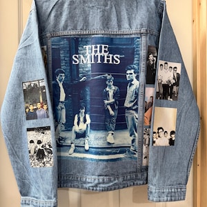 May include: Light blue denim jacket with a large graphic of The Smiths on the back. The jacket features several smaller black and white photo patches of the band. It has a button-up front and a collar.