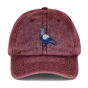 Peut inclure: Une casquette de baseball bordeaux avec une finition délavée à l'acide. Le panneau avant présente un motif brodé représentant un pigeon au plumage bleu, gris et blanc. La casquette possède une visière courbée, un bouton et des œillets.
