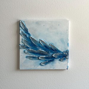 Könnte beinhalten: Quadratische Leinwandkunst mit einem strukturierten, erhabenen Design. Das Kunstwerk zeigt eine blau-weiße Farbpalette mit einer stilisierten, flügelförmigen Form in Blau- und Silbertönen. Der Hintergrund ist ein weiches, helles Blau.