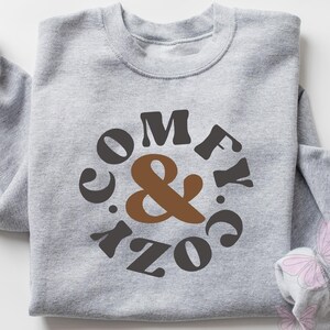 May include: A light gray sweatshirt with the words "COMFY & COZY" in a circular design. The text is in a mix of brown and dark gray. The sleeves are rolled up. Pink butterflies are in the background.