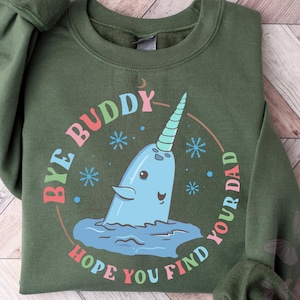 Pode incluir: Um moletom verde-oliva com o texto "Bye Buddy Hope You Find Your Dad" em um design circular. O design apresenta uma narval azul com um chifre arco-íris, cercado por flocos de neve e uma lua crescente.