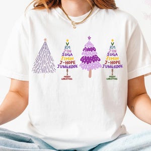 Op de afbeelding: Wit T-shirt met drie kerstboomontwerpen in paars en groen. Elke boom heeft de tekst "RM, Jin, Suga, Jimin, J-Hope, Jungkook" en "Merry Christmas".