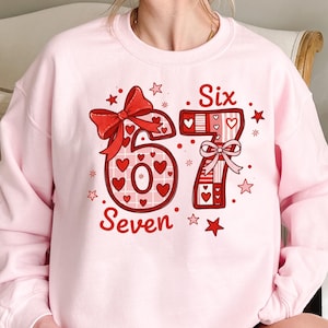 May include: A light pink sweatshirt with a red graphic design. The design features the numbers "6" and "7" with hearts, bows, and stars, and the words "Six" and "Seven".