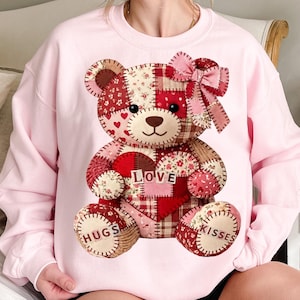 Könnte beinhalten: Ein hellrosa Sweatshirt mit einem Patchwork-Teddybär-Design. Der Bär hat ein Herz mit dem Wort "LOVE" und den Worten "HUGS" und "KISSES" auf seinen Pfoten. Der Bär ist mit einer rosa Schleife verziert.