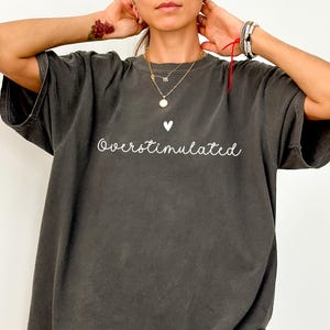 May include: Dark gray oversized t-shirt with the word "Overstimulated" in white script, above a small white heart. The shirt has short sleeves and a relaxed fit, perfect for casual wear.
