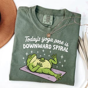 May include: Green t-shirt with the text "Today's yoga pose is DOWNWARD SPIRAL" and a cartoon frog on a yoga mat. The shirt has a crew neck and short sleeves. A gold necklace is draped across the shirt.