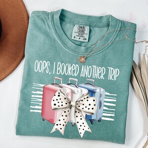 May include: A teal Comfort Colors t-shirt with the phrase "oops, I booked another trip" printed on the front. The graphic features three suitcases and a large bow. A gold necklace is draped across the shirt.