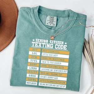 Puede incluir: Una camiseta Comfort Colors verde mar con el texto "SENIOR CITIZEN TEXTING CODE" y una lista de acrónimos de mensajes de texto. Un collar dorado con un colgante está sobre la camiseta.