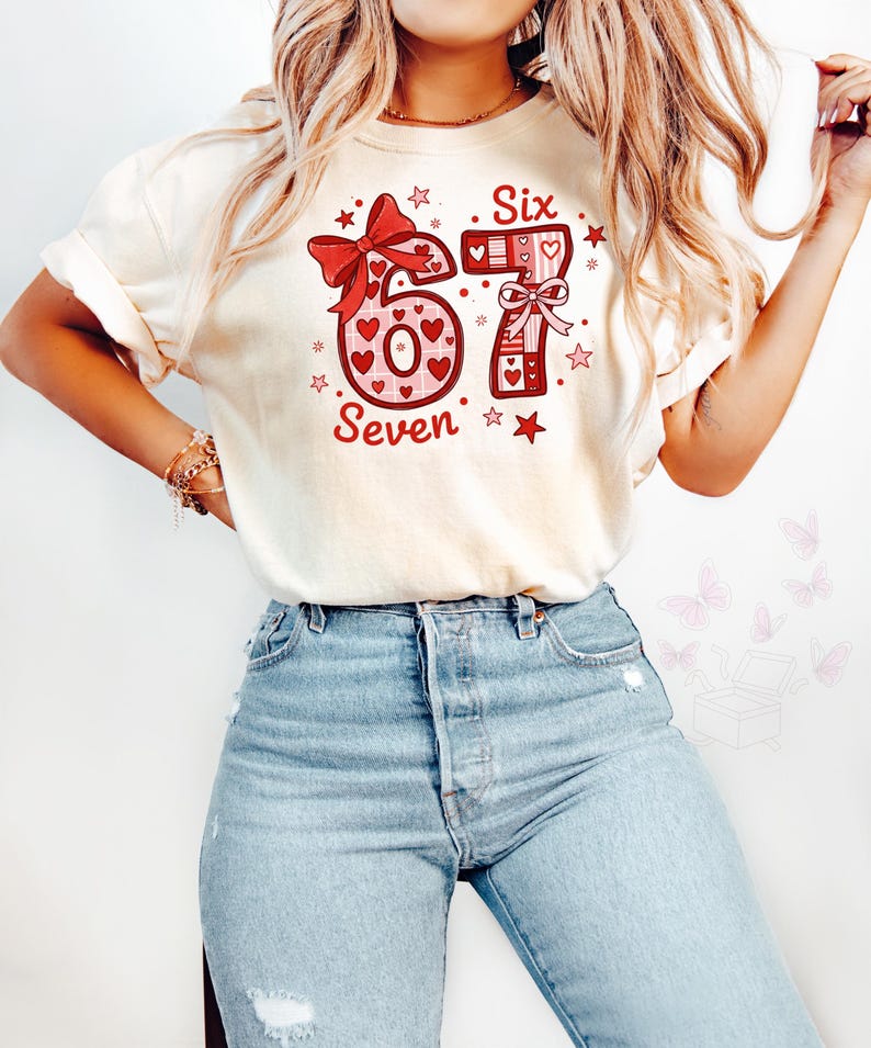 May include: Cream-colored t-shirt with a large graphic featuring the numbers "6" and "7" in red, decorated with hearts and bows. The word "Six" is above the "6", and "Seven" is below. Stars and hearts surround the numbers.
