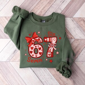 May include: A green sweatshirt with the numbers "6" and "7" in a red and white design. The number "6" has a bow and heart accents. The number "7" has a bow and heart accents. The word "Seven" is below the number "6". The word "Six" is above the number "7".