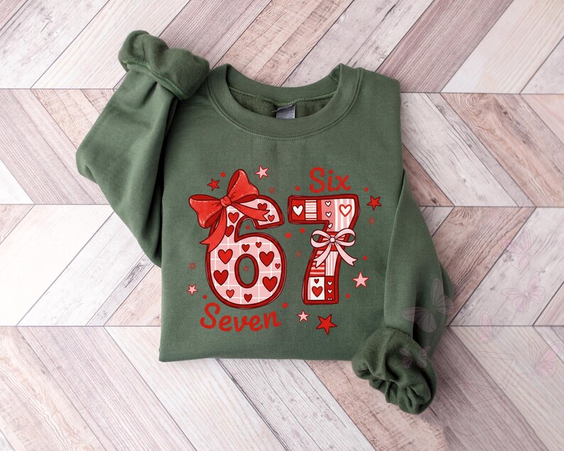 May include: A green sweatshirt with the numbers "6" and "7" in a red and white design. The number "6" has a bow and heart accents. The number "7" has a bow and heart accents. The word "Seven" is below the number "6". The word "Six" is above the number "7".