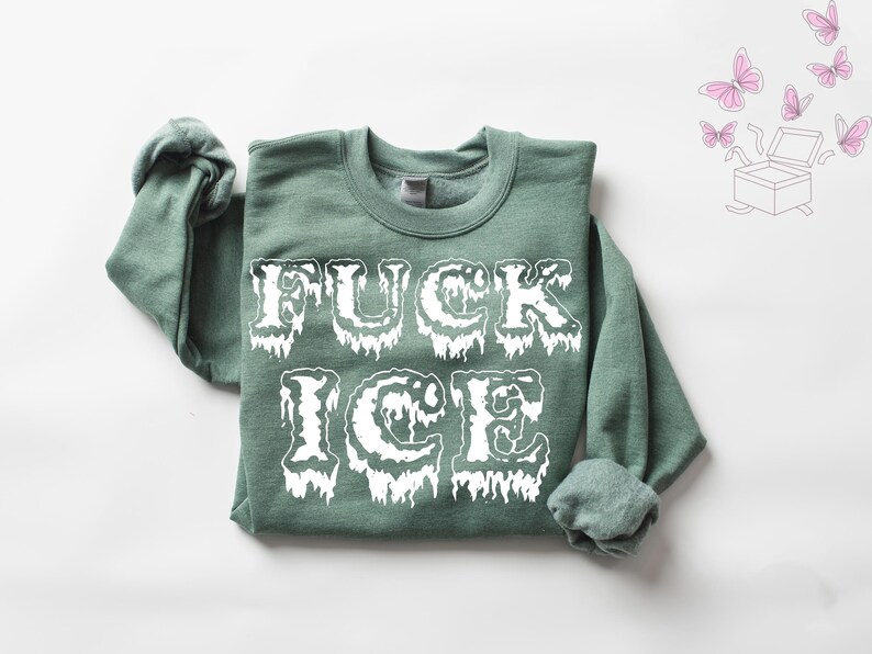 Pu&ograve; includere: Una felpa verde salvia con la scritta "FUCK ICE" in grandi lettere bianche gocciolanti. Le maniche sono arrotolate. Sullo sfondo ci sono farfalle rosa e un'illustrazione di una scatola.