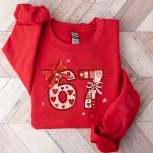 May include: Red sweatshirt with a graphic design featuring the number "67" decorated with hearts and bows. The word "Seven" is printed below the numbers. The sleeve has a butterfly design.