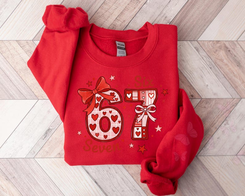 May include: Red sweatshirt with a graphic design featuring the number "67" decorated with hearts and bows. The word "Seven" is printed below the numbers. The sleeve has a butterfly design.
