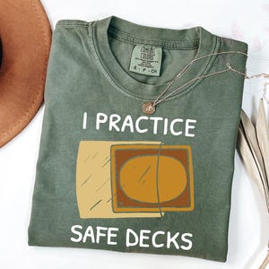 May include: Olive green t-shirt with the text "I PRACTICE SAFE DECKS" and a graphic of a card. A gold necklace is draped over the shirt. The shirt is folded on a white surface.