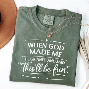 May include: An olive green t-shirt with the text "WHEN GOD MADE ME HE GRINNED AND SAID 'This'll be fun'" in white. A gold necklace with a circular pendant rests on the shirt. A brown hat is visible in the upper left corner.