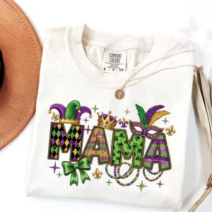 以下が含まれることがあります： クリーム色のTシャツに、カラフルなマルディグラのデザインで「MAMA」の文字。道化師の帽子、王冠、マスク、ビーズのデザイン。ペンダント付きのゴールドネックレスがシャツの上にかけられています。