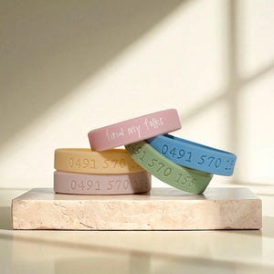 May include: A collection of pastel-coloured silicone wristbands. The pink band reads "find my folks". Other bands display a phone number. The bands are stacked on a small, square, light-coloured block. The wristbands are in shades of pink, yellow, blue, and green.