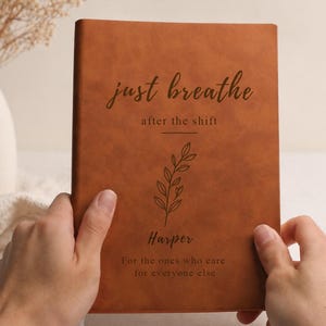 Könnte beinhalten: Ein braunes Lederjournal mit den Worten "just breathe" und "after the shift" in eleganter Schrift. Unter dem Text befindet sich ein floraler Zweig. Der Name "Harper" ist unter dem Zweig aufgedruckt, mit dem Satz "For the ones who care for everyone else" darunter.