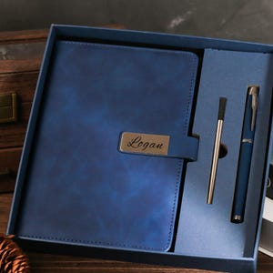 Könnte beinhalten: Ein blaues Leder-Notizbuch- und Stift-Set in einer Geschenkbox. Das Notizbuch hat ein Metallschild mit dem eingravierten Namen "Logan". Das Set enthält einen passenden Stift und einen silbernen Stift. Die Box ist blau.