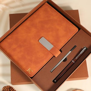 May include: A brown leather journal with a silver metal clasp and the initials "JC" in gold, presented in a brown gift box. Includes a silver pen and a dark brown pen. A thoughtful gift set.
