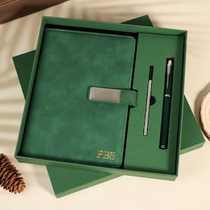 Può includere: Un set regalo con diario e penna in pelle verde. Il diario ha una chiusura argentata e le iniziali "PBS" in oro. La penna e il diario sono verde scuro.