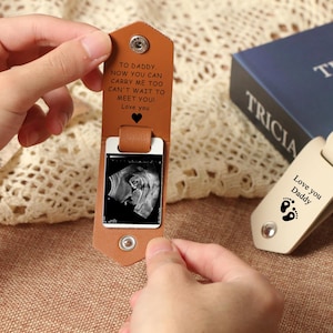 May include: A brown leather keychain with a silver frame displaying a sonogram image. The keychain features the text "TO DADDY, NOW YOU CAN CARRY ME TOO CAN'T WAIT TO MEET YOU! Love you" and a heart. Another keychain says "Love you Daddy" with footprints.