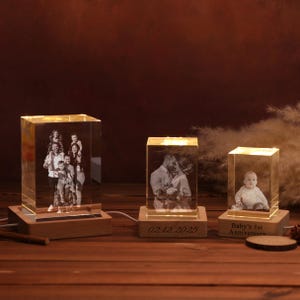 Peut inclure: Trois blocs photo en cristal éclairés avec des portraits de famille. Chaque bloc repose sur une base en bois. Le bloc de droite porte l'inscription "Baby's 1st Anniversary". Le bloc du milieu porte la date "02.12.2025".