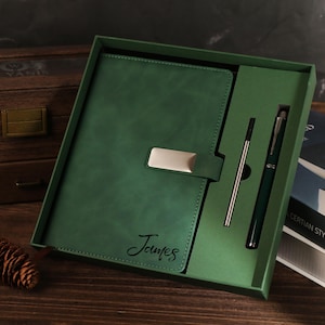 Könnte beinhalten: Ein grünes Leder-Notizbuch- und Stift-Set in einer Geschenkbox. Das Notizbuch hat einen silbernen Verschluss und den Namen "James" auf dem Einband. Der Stift ist dunkelgrün mit silbernen Akzenten. Das Set liegt auf einer Holzoberfläche.