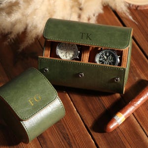 May include: Green leather watch cases, one open with two watches inside, and a closed single watch case. The open case has the initials "TK" on the lid. A cigar rests on a wooden surface.