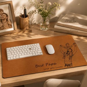 Op de afbeelding: Een bruine leren bureaumat met een wit toetsenbord en muis. De mat heeft een lijntekening van een vader en kind en de tekst "Best Papa Love you Lucas". Een ingelijste foto en boeken staan ook op het bureau.