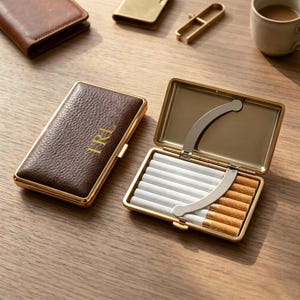 Könnte beinhalten: Ein rechteckiges Zigarettenetui mit einer dunkelbraunen, genarbten Lederoberfläche und den gold geprägten Buchstaben TRL. Das Etui hat einen goldfarbenen Metallrahmen und ein offenes Inneres, in dem mehrere Zigaretten von einem Metallbügel gehalten werden.
