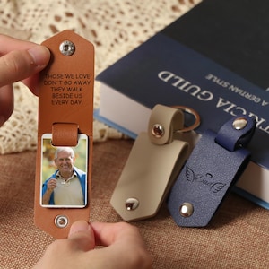 May include: Leather keyrings in tan and navy blue, with a photo and text. One keyring has a photo of a man, and the other has the word "Dad" with angel wings. The text on the first keyring reads "Those we love don't go away, they walk beside us every day."