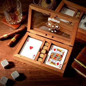 May include: A wooden box with a glass lid featuring the name Benjamin and card suit icons. Inside are two decks of playing cards and five gold-coloured dice. Beside the box are two cigars, a glass of amber liquid, and three grey stone ice cubes.